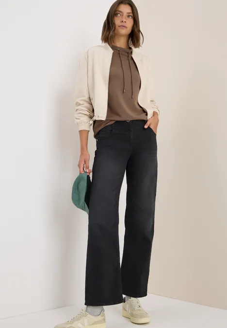 Frau in weiter Jeans, braunem Hoodie und beiger Jacke hält eine grüne Kappe.